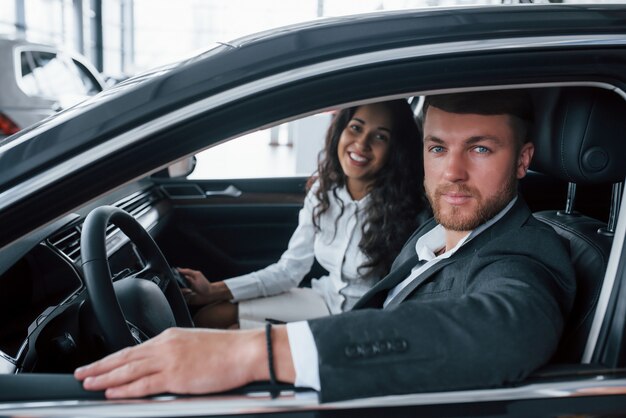 Retrato de familia en el vehículo. Encantadora pareja exitosa probando coche nuevo en el salón del automóvil