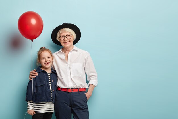 El retrato familiar de la nieta y la abuela se abrazan y celebran las vacaciones, sostienen el globo de aire, usan ropa festiva, expresan emociones positivas aisladas en la pared azul. Concepto de generación y fiesta