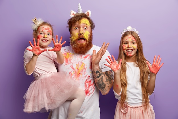 El retrato familiar del padre sorprendido tiene la cara amarilla de las pinturas, dos hijas alegres muestran palmas sucias con acuarelas, cuadro de pintura durante el pasatiempo, aislado en la pared lila. Esto es arte