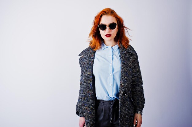 Retrato de una fantástica chica pelirroja con camisa azul y abrigo gris posando con gafas de sol en el estudio