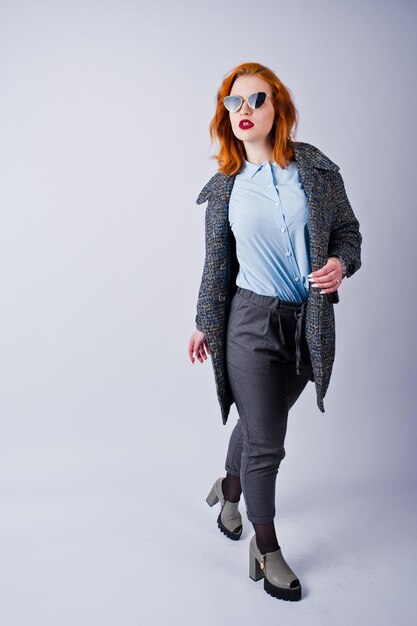 Retrato de una fantástica chica pelirroja con camisa azul y abrigo gris posando con gafas de sol en el estudio