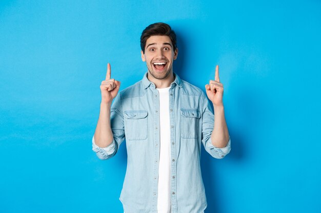 Retrato de feliz 25 años chico con barba, apuntando con el dedo hacia arriba y sonriendo, mostrando publicidad, de pie contra el fondo azul.