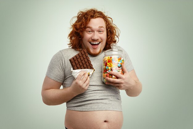 Retrato de un feliz y alegre hombre pelirrojo con una camiseta de talla pequeña de pie, disfrutando de dulces, mermeladas y chocolate poco saludables pero deliciosos