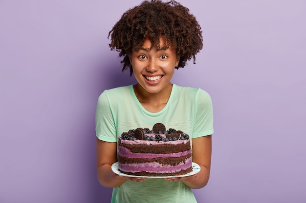 Retrato de feliz ama de casa de piel oscura demuestra un delicioso pastel horneado por ella misma, quiere tratar a los invitados, tiene buenas habilidades coolinary