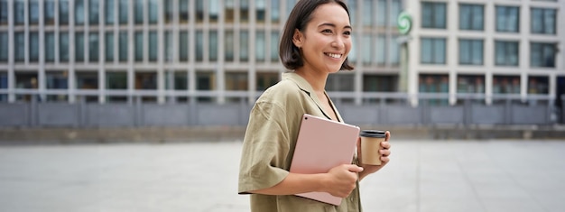 Foto gratuita retrato de una feliz chica asiática con tableta y café caminando por la calle con una sonrisa complacida