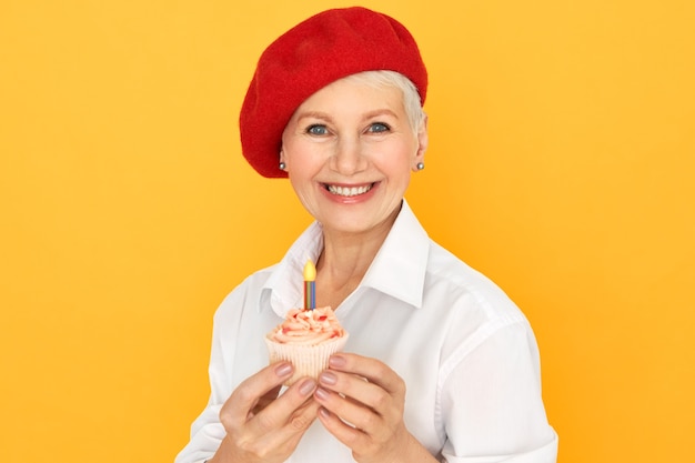 Retrato de feliz encantadora mujer caucásica de mediana edad en elegante sombrero rojo celebrando su cumpleaños, posando aislado con cupcake en sus manos. Concepto de celebración, fiesta y ocasiones especiales.