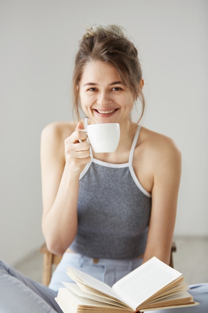 Retrato de feliz hermosa joven sonriente hoding taza de café y libro sentado en el piso sobre la pared blanca