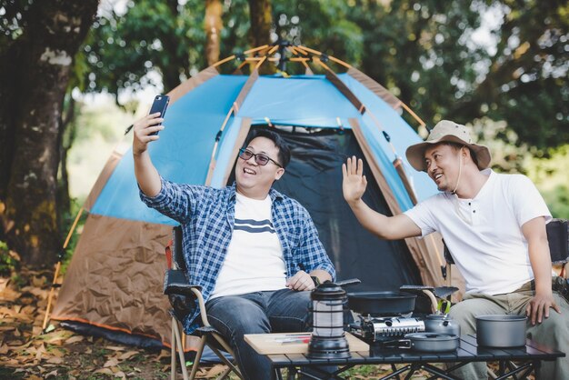 Retrato feliz hombre asiático amigos Hacer una videollamada con un teléfono inteligente en el campamento Juego de cocina en el frente Cocina al aire libre Viajar acampar concepto de estilo de vida