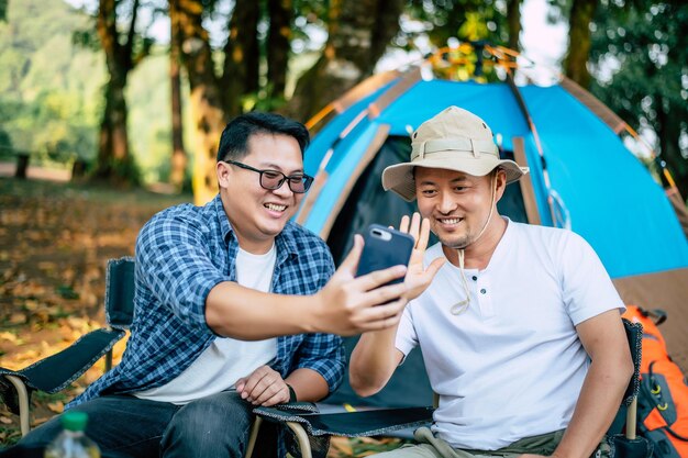 Retrato feliz hombre asiático amigos Hacer una videollamada con un teléfono inteligente en el campamento Juego de cocina en el frente Cocina al aire libre Viajar acampar concepto de estilo de vida