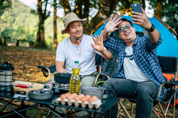 Retrato feliz hombre asiático amigos Hacer una videollamada con un teléfono inteligente en el campamento Juego de cocina en el frente Cocina al aire libre Viajar acampar concepto de estilo de vida