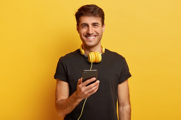 Retrato de feliz hombre caucásico disfruta de un sonido fantástico y la calidad de los auriculares, sostiene un teléfono celular moderno