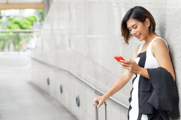 Retrato feliz joven y bella mujer de pie cerca de la pared sonriendo y uso smartphone