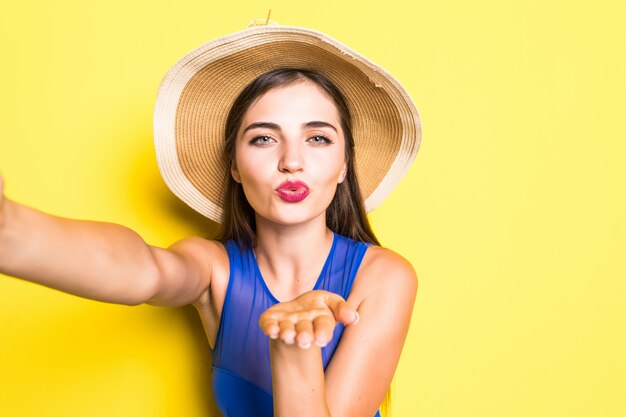 Retrato de feliz joven y bella mujer tomando selfie en su teléfono y sonriendo, vistiendo traje de baño con sombrero de paja en la pared amarilla