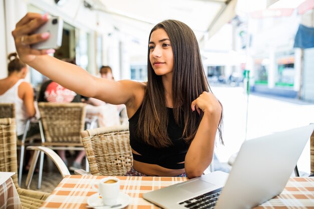 Retrato de feliz joven latina tomando selfie con teléfono móvil mientras está sentado en un café al aire libre