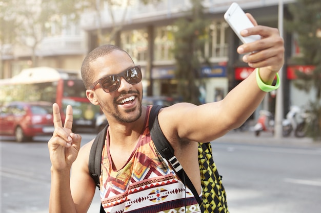 Retrato de feliz joven de piel oscura en gafas de sol y camiseta sin mangas sonriendo mientras toma selfie posando con gesto de paz