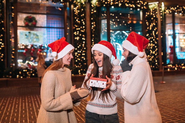 Retrato de feliz lindo joven grupo de amigos abrazándose y sonriendo mientras camina en la víspera de Navidad al aire libre, con sombreros de santa s, muchas luces en el fondo