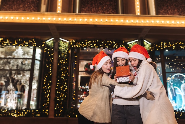 Foto gratuita retrato de feliz lindo joven grupo de amigos abrazándose y sonriendo mientras camina en la víspera de navidad al aire libre, con sombreros de santa s, muchas luces en el fondo