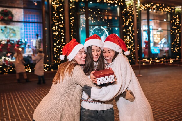 Retrato de feliz lindo joven grupo de amigos abrazándose y sonriendo mientras camina en la víspera de Navidad al aire libre, vistiendo gorros de santa, muchas luces en el