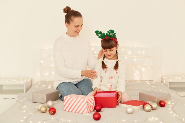 Retrato de feliz madre e hija abriendo regalos de Navidad en la cama en casa, pasando la víspera de año nuevo juntos, celebrando las vacaciones de invierno, feliz Navidad.