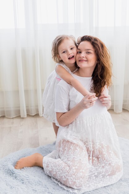 Retrato de feliz madre e hija sentada en la suave alfombra mirando a la cámara