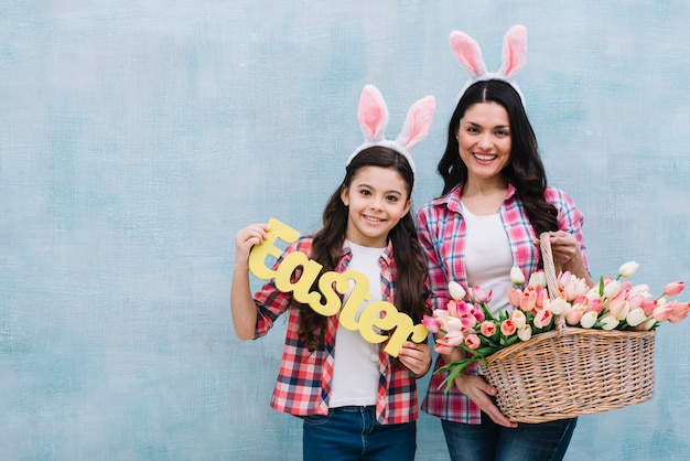 Retrato feliz de la madre y la hija que sostienen la palabra de pascua y la cesta de los tulipanes contra la pared azul