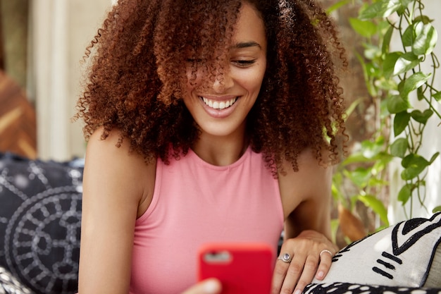 Retrato de feliz modelo femenino multiétnico mira películas en línea en un teléfono inteligente moderno, conectado a internet inalámbrico, se sienta en un cómodo sofá. mujer joven atractiva lee noticias de la red social