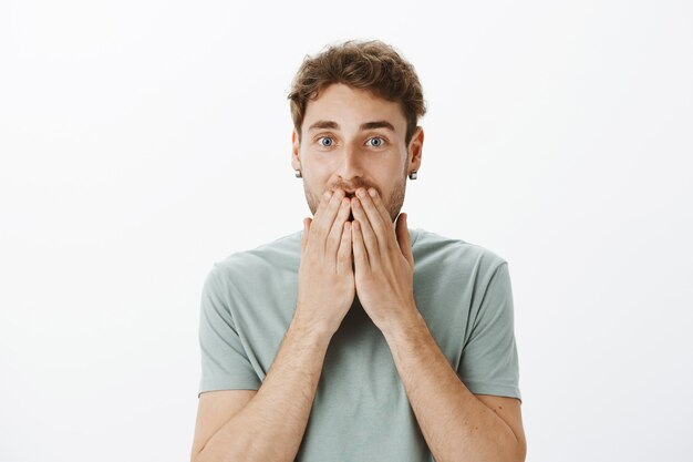 Retrato de feliz modelo masculino europeo en aretes, sonriendo y cubriendo la boca con las palmas, sorprendido y satisfecho con una gran noticia