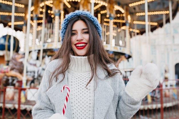 Retrato de feliz modelo de mujer de pelo largo en guantes blancos con bastón de caramelo en el parque de atracciones. Chica guapa con sombrero azul celebrando la Navidad y posando junto al carrusel.