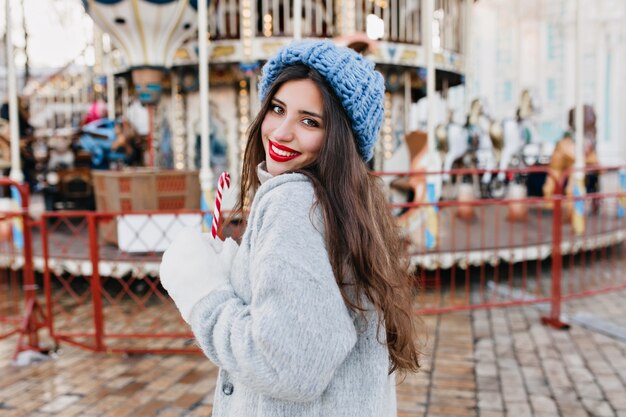 Retrato de feliz modelo de mujer de pelo largo en guantes blancos con bastón de caramelo en el parque de atracciones. Chica guapa con sombrero azul celebrando la Navidad y posando junto al carrusel.