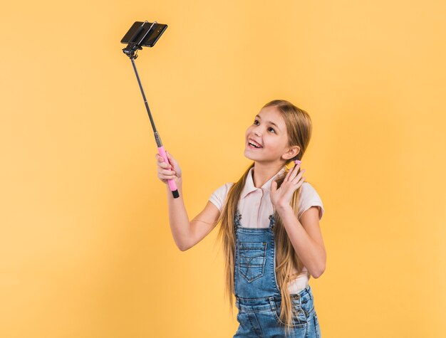 Retrato feliz de una muchacha que agita su mano que toma el selfie en el teléfono móvil contra fondo amarillo