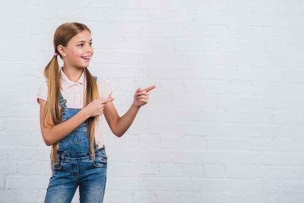 Foto gratuita retrato feliz de una muchacha que señala el dedo que se opone a la pared de ladrillo blanca que mira lejos