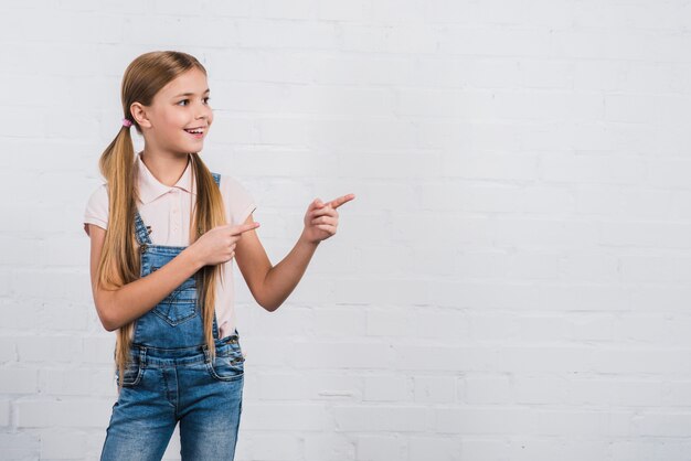 Retrato feliz de una muchacha que señala el dedo que se opone a la pared de ladrillo blanca que mira lejos