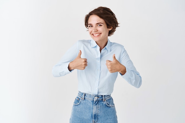 Retrato de feliz mujer caucásica muestra los pulgares hacia arriba y sonríe en aprobación, elogia el buen trabajo, el gesto bien hecho, me gusta y recomienda el producto, de pie en la pared blanca