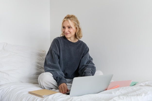 Retrato de una feliz mujer rubia independiente que trabaja desde casa sentada en la cama con una computadora portátil y cuadernos