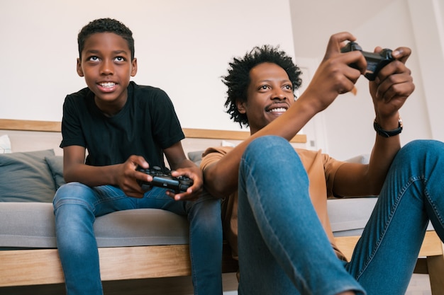 Retrato de feliz padre e hijo afroamericano sentados en el sofá cama y jugando videojuegos de consola juntos en casa. Concepto de familia y tecnología.