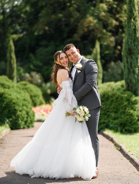 Retrato de feliz pareja casada guapo novio en elegante traje gris abrazando a la novia en vestido de novia con ramo mientras posa en el jardín Ceremonia del día de la boda