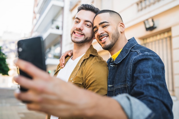 Retrato de feliz pareja gay pasar tiempo juntos y tomar un selfie con teléfono móvil en la calle.