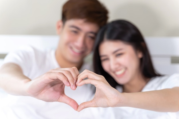 Retrato feliz pareja joven sentada y sonriendo en la cama y la mano hacen juntos la forma del corazón