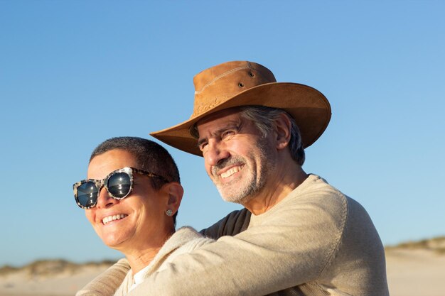 Retrato de feliz pareja mayor pasando tiempo juntos al aire libre en un día soleado. Un hombre canoso sonriente con sombrero de vaquero abrazando a una mujer feliz de pelo corto con gafas de sol. Amor, concepto de ocio