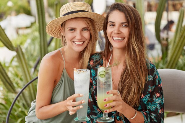 Retrato de feliz pareja de mujeres homosexuales disfrutar del descanso de verano, celebrar algo en un acogedor restaurante, tintinear vasos de cócteles, tener amplias sonrisas. Encantadora mujer joven con sombrero con un amigo cercano en la barra