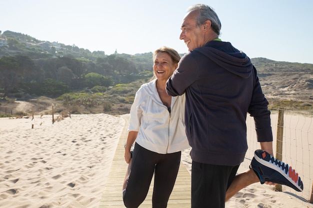 Retrato de feliz pareja senior haciendo ejercicios físicos. Mujer y hombre sonrientes parados cerca al aire libre estirando las piernas tratando de mantenerse en forma y saludables. Deporte, estilo de vida saludable del concepto de personas mayores