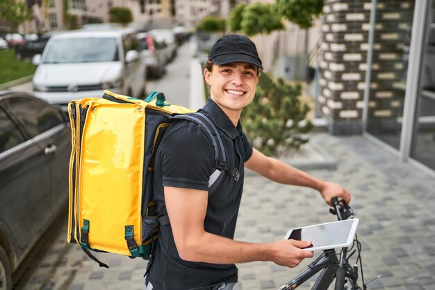 Retrato de feliz repartidor en uniforme en bicicleta cerca de una casa moderna, con una mochila amarilla