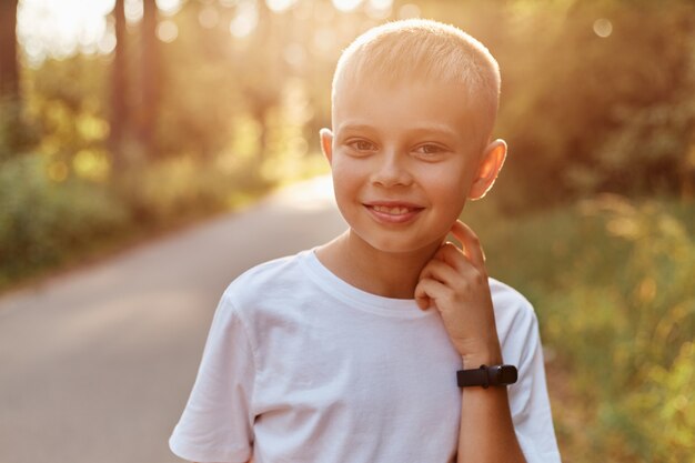 Retrato de feliz sonriente niño rubio con camiseta blanca casual, mirando directamente a la cámara con una gran sonrisa, manteniendo la mano en el cuello, pasando tiempo en el parque de verano al atardecer.