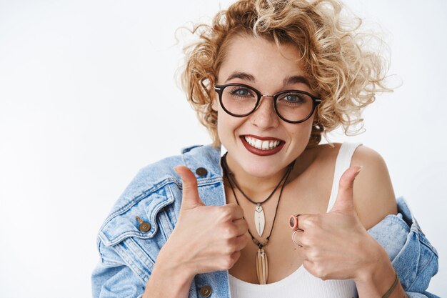 Retrato de feliz vítores amigable y solidaria mujer joven complacida con gafas y chaqueta de mezclilla sonriendo felizmente mostrando los pulgares para arriba gesto en me gusta y aprobación