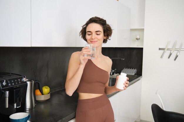 Retrato de fitness chica mujer sana de pie en la cocina con pastillas toma vitaminas con vaso de w