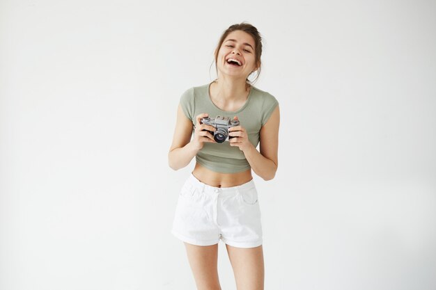 Retrato de la fotografía femenina joven feliz alegre que ríe sosteniendo la cámara vieja sobre la pared blanca.