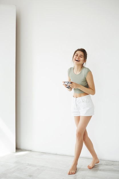 Retrato de la fotografía femenina joven feliz alegre que ríe sosteniendo la cámara vieja sobre la pared blanca.