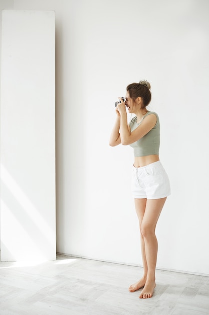 Retrato de la fotografía femenina joven feliz alegre que sonríe tomando la imagen en cámara vieja sobre la pared blanca.