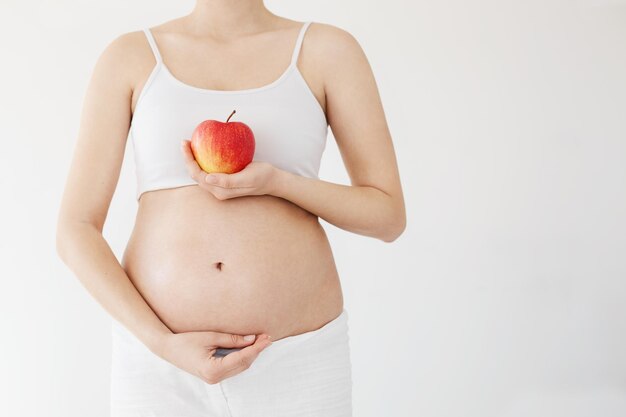 Retrato frontal de una mujer embarazada sana sosteniendo su vientre y una manzana roja llena de vitaminas Concepto de dieta saludable para el embarazo sobre blanco Clave alta