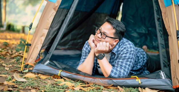 Retrato de gafas de hombre viajero asiático feliz acostado en una tienda de campaña acampar al aire libre y concepto de estilo de vida